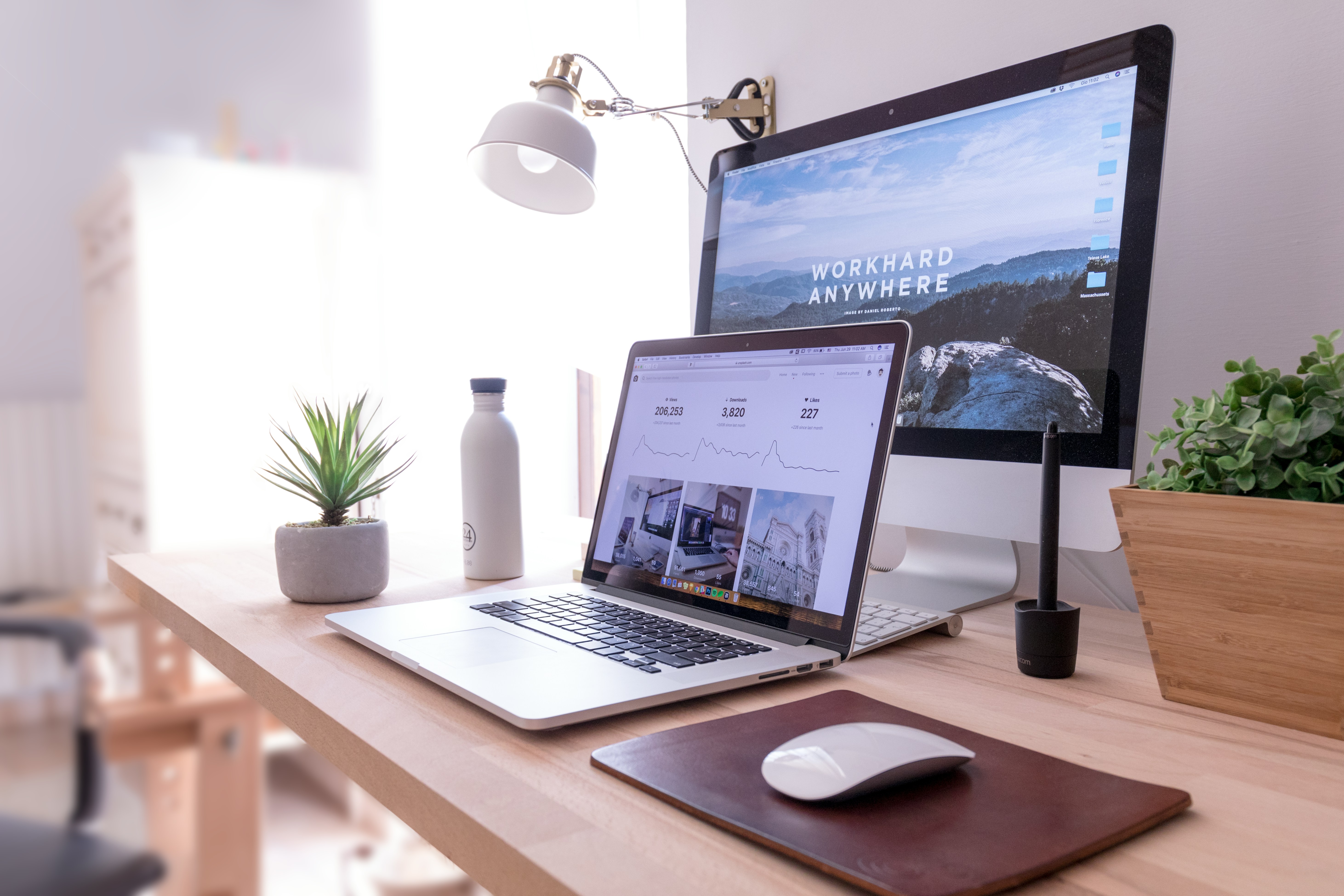 Bureau avec un ordinateur portable et un écran affichant des interfaces web, accompagnés d’une souris, d’une lampe et de plantes décoratives.