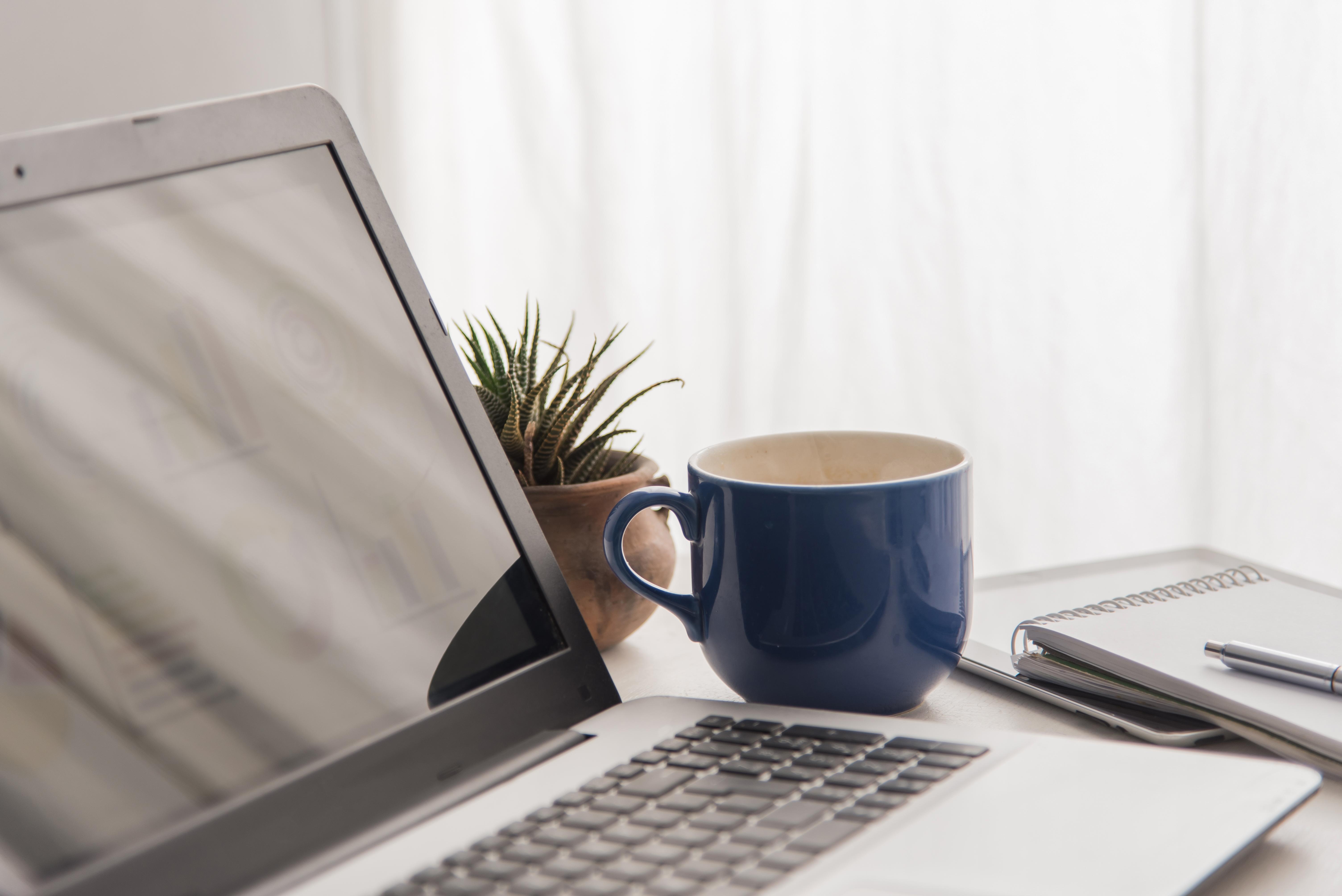 Ordinateur portable ouvert sur un bureau, accompagné d’une tasse de café bleu, d’une plante et de carnets de notes.