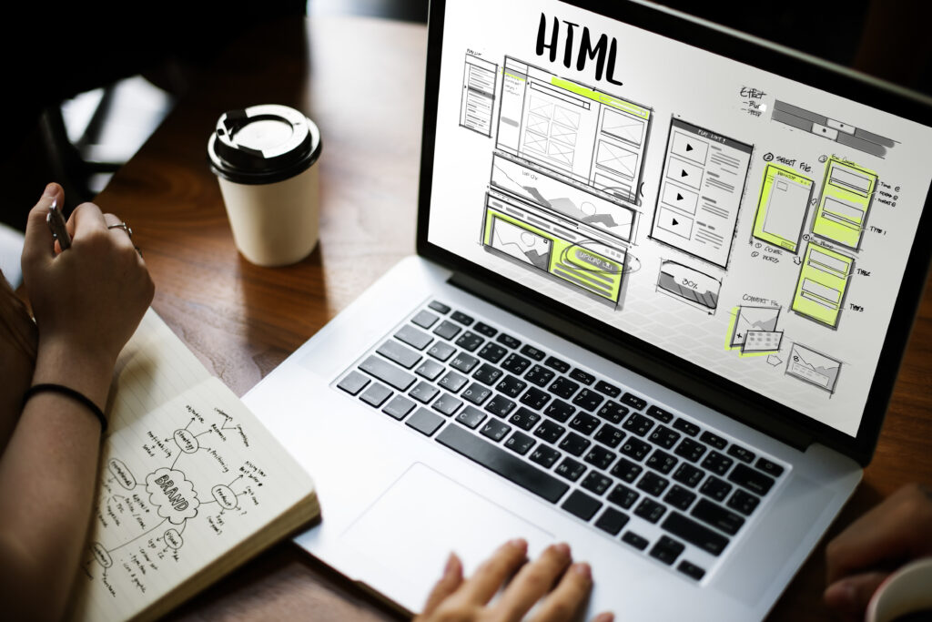 Personne travaillant sur la conception d’un site web HTML avec un ordinateur portable, un carnet de croquis et une tasse de café sur un bureau.